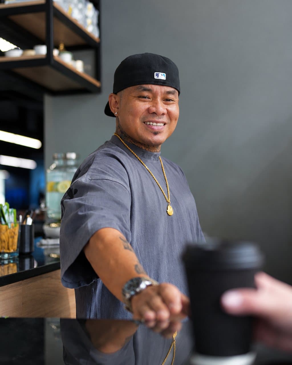 Someone in a black cap smiling behind a coffee bar, reaching out a fist bump to a hand holding a takeaway coffee